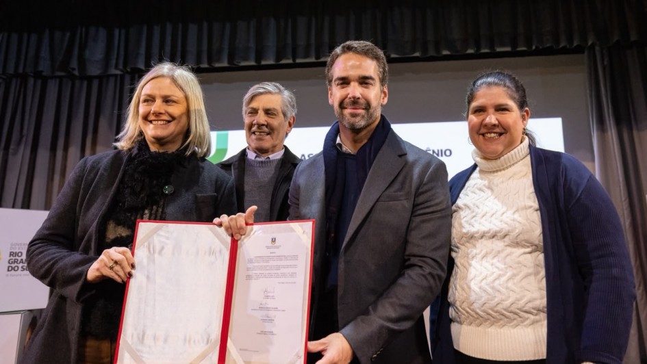 **Descri&ccedil;&atilde;o da imagem "Governo do Estado e munic&iacute;pio de Pelotas assinam conv&ecirc;nio do Programa Iconicidades para implanta&ccedil;&atilde;o de Centro de Gastronomia.jpeg":**

Uma fotografia colorida em plano m&eacute;dio e vis&atilde;o frontal mostra quatro pessoas sorrindo no palco de um audit&oacute;rio, posando para a foto enquanto exibem um documento oficial assinado.

**Os Participantes (da esquerda para a direita):**

1. Uma mulher de pele clara e cabelos loiros curtos, na altura dos ombros. Ela sorri olhando levemente para a esquerda e segura a borda esquerda de uma pasta de capa vermelha aberta. Veste um casaco sobretudo preto com um cachecol felpudo preto e cal&ccedil;a marrom.
2. Um homem mais velho de cabelos grisalhos, posicionado um pouco mais ao fundo. Ele sorri olhando na mesma dire&ccedil;&atilde;o que a primeira mulher e veste um casaco preto sobre um su&eacute;ter cinza de gola careca.
3. O governador Eduardo Leite, posicionado no centro-direito. Ele tem barba curta aparada, sorri diretamente para a c&acirc;mera e segura a borda inferior da pasta vermelha aberta. Veste um palet&oacute; cinza-escuro sobre uma blusa azul-marinho com um cachecol azul-escuro no pesco&ccedil;o.
4. Uma mulher de cabelos escuros presos em um rabo de cavalo. Ela sorri de frente para a c&acirc;mera e usa um cardig&atilde; azul-marinho aberto sobre uma blusa de tric&ocirc; branca de gola alta trabalhada com texturas, al&eacute;m de brincos dourados pendentes.

**O Documento e o Cen&aacute;rio:**

* No centro do grupo, a pasta de capa vermelha est&aacute; aberta e voltada para a frente, revelando em sua p&aacute;gina direita um documento oficial impresso em papel branco com o bras&atilde;o do Estado no topo e assinaturas com carimbos na parte inferior. A p&aacute;gina esquerda da pasta est&aacute; em branco.
* Ao fundo, uma tela de proje&ccedil;&atilde;o clara exibe parcialmente a palavra **"V&Ecirc;NIO"** em letras escuras e mai&uacute;sculas.
* Na parte superior e nas laterais do palco, h&aacute; cortinas e franjas de tecido cinza-escuro dispostas verticalmente. No canto extremo esquerdo, avista-se um detalhe de um totem branco com o logotipo oficial do **"GOVERNO DO ESTADO RIO GRANDE DO SUL"**. A ilumina&ccedil;&atilde;o &eacute; frontal e focada no grupo.