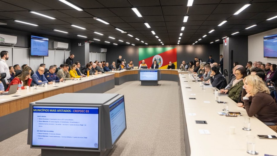 Grande sala com v&aacute;rias pessoas sentadas junto a uma grande mesa oval, com a bandeira do Rio Grande do Sul ao fundo e paineis no centro da mesa.