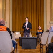 **Descri&ccedil;&atilde;o da imagem "Governo do Estado alerta para riscos associados &agrave;s chuvas intensas nos pr&oacute;ximos dias.jpeg":**

Uma fotografia colorida em plano m&eacute;dio e enquadramento centralizado mostra pronunciamento oficial conduzido no interior de um sal&atilde;o nobre ou audit&oacute;rio cl&aacute;ssico.

**O Orador Principal:**
No centro da imagem, o governador Eduardo Leite est&aacute; em p&eacute; diante de uma cortina dourada. Ele tem barba curta aparada, segura um microfone com a m&atilde;o direita perto da boca e gesticula levemente com a m&atilde;o esquerda enquanto fala. Veste um palet&oacute; azul-marinho ou preto sobre uma camisa social branca sem gravata e cal&ccedil;a social cinza.

**Outros Participantes e Plateia:**

* &Agrave; direita de Eduardo Leite, sentado em uma cadeira clara ao fundo, est&aacute; um agente da Defesa Civil de cabe&ccedil;a calva, vestindo uma jaqueta operacional azul e preta com faixa refletiva e detalhes em laranja no peito, al&eacute;m de cal&ccedil;a escura.
* Em primeiro plano, vistos de costas e de perfil, h&aacute; integrantes da plateia sentados em cadeiras estofadas em tom bege/creme. &Agrave; esquerda, destaca-se um homem de costas usando um colete operacional laranja com a inscri&ccedil;&atilde;o em azul **"DEFESA CIVIL ALVORADA RS"**.
* No canto esquerdo, avista-se parte de um painel azul onde se l&ecirc; em letras mai&uacute;sculas brancas **"CLIM&Aacute;TICOS E PREVENTIV..."**.

**O Ambiente:**

* O espa&ccedil;o possui arquitetura cl&aacute;ssica e elegante, com paredes em tom creme/amarelo-claro, colunas decorativas e dois lustres de estilo vintage acesos nas paredes laterais (um de cada lado da cortina).
* Ao fundo, uma imponente e alta cortina de tecido dourado/mostarda cobre toda a extens&atilde;o central da parede.
* &Agrave; frente do governador, no ch&atilde;o, h&aacute; um monitor de retorno retangular preto voltado para a plateia. O piso do sal&atilde;o &eacute; de madeira em trabalho de parquet estruturado em formas geom&eacute;tricas.