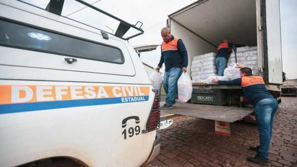 

Uma fotografia colorida em plano m&eacute;dio e perspectiva em contra-plong&eacute;e (de baixo para cima) mostra uma opera&ccedil;&atilde;o de descarregamento de doa&ccedil;&otilde;es ou suprimentos de ajuda humanit&aacute;ria realizada por agentes da Defesa Civil.

**Os Agentes e a A&ccedil;&atilde;o:**

* Tr&ecirc;s homens vestindo o uniforme operacional da Defesa Civil &mdash; camisetas de mangas compridas em azul-marinho com ombros e gola em laranja, e cal&ccedil;as jeans &mdash; participam do trabalho.
* No centro, um homem caminha sobre a plataforma elevat&oacute;ria traseira do caminh&atilde;o, carregando dois grandes sacos pl&aacute;sticos transparentes cheios de suprimentos, um em cada m&atilde;o.
* &Agrave; direita, em primeiro plano, outro agente est&aacute; em p&eacute; na rua, esticando os bra&ccedil;os para receber um pacote do interior do ve&iacute;culo.
* Mais ao fundo, no interior do ba&uacute; do caminh&atilde;o, um terceiro agente organiza fardos empilhados de doa&ccedil;&otilde;es mantidas em embalagens pl&aacute;sticas transparentes.

**Os Ve&iacute;culos e o Ambiente:**

* No canto esquerdo, em primeiro plano, destaca-se a lateral traseira de uma caminhonete branca da Defesa Civil. Na lataria, h&aacute; uma faixa horizontal laranja e azul com a inscri&ccedil;&atilde;o em letras mai&uacute;sculas **"DEFESA CIVIL ESTADUAL"** e o n&uacute;mero de emerg&ecirc;ncia **"199"** acompanhado do &iacute;cone de um telefone em preto. No teto do ve&iacute;culo, v&ecirc;-se um bagageiro de metal preto.
* &Agrave; direita, o caminh&atilde;o-ba&uacute; de carga est&aacute; com as portas traseiras totalmente abertas e a plataforma hidr&aacute;ulica abaixada ao n&iacute;vel de acesso.
* O solo &eacute; de pavimento de paralelep&iacute;pedos em tons avermelhados e o c&eacute;u ao fundo est&aacute; nublado e claro.