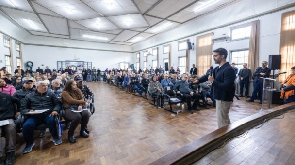 Um homem fala ao microfone diante de uma grande plateia reunida em um sal&atilde;o amplo. H&aacute; dezenas de pessoas sentadas em cadeiras e outras em p&eacute; ao fundo.