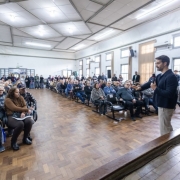 Um homem fala ao microfone diante de uma grande plateia reunida em um sal&atilde;o amplo. H&aacute; dezenas de pessoas sentadas em cadeiras e outras em p&eacute; ao fundo.