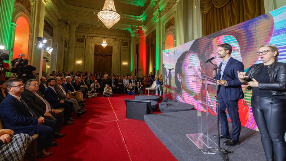 Foto de sal&atilde;o de estilo cl&aacute;ssico, com tapete vermelho. Um homem fala ao p&uacute;lpito no canto direito, em trajes formais. A plateia est&aacute; cheia e ocupa o lado esquerdo e o fundo do quadro, com a maior parte das pessoas sentada.