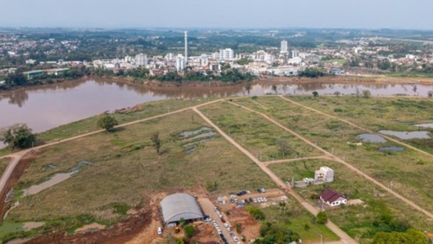 Uma fotografia a&eacute;rea colorida em plano aberto e &acirc;ngulo picado (tomada por drone) mostra uma ampla extens&atilde;o de terreno descampado cortado por um rio, com uma &aacute;rea urbana vis&iacute;vel ao fundo.

**O Terreno e as Edifica&ccedil;&otilde;es em Primeiro Plano:**

* Em primeiro plano e ao centro, estende-se uma vasta &aacute;rea descampada de topografia plana, coberta por vegeta&ccedil;&atilde;o rasteira verde e seca, entremeada por alguns po&ccedil;os d&rsquo;&aacute;gua e pequenas &aacute;reas alagadas.
* O terreno &eacute; recortado por tra&ccedil;ados de vias de terra batida clara, delineando futuros acessos ou quadras.
* Na parte inferior central, h&aacute; uma estrutura tipo galp&atilde;o com cobertura curva em arco met&aacute;lico cinza. Ao lado e ao redor do galp&atilde;o, v&ecirc;-se uma concentra&ccedil;&atilde;o de dezenas de ve&iacute;culos (carros de passeio e vans) estacionados em fileiras sobre a terra batida, indicando a realiza&ccedil;&atilde;o de um evento no local.
* Pr&oacute;ximo ao galp&atilde;o e ao longo da borda inferior direita, observam-se algumas casas residenciais de telhado colonial cercadas por &aacute;rvores e ruas de acesso.

**O Rio e o Entorno Urbano ao Fundo:**

* Logo acima do grande terreno, uma curva larga de rio com &aacute;guas barrentas e amarronzadas atravessa horizontalmente a paisagem.
* Na margem oposta do rio, estende-se a mancha urbana do munic&iacute;pio, caracterizada por diversos pr&eacute;dios brancos de m&eacute;dio e grande porte, edifica&ccedil;&otilde;es comerciais, resid&ecirc;ncias e uma alta chamin&eacute; ou torre branca cil&iacute;ndrica centralizada.
* Ao fundo da cidade, a paisagem se desdobra em colinas suaves com vegeta&ccedil;&atilde;o densa sob um c&eacute;u azul-claro com nuvens difusas na linha do horizonte.