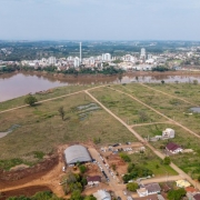 Uma fotografia a&eacute;rea colorida em plano aberto e &acirc;ngulo picado (tomada por drone) mostra uma ampla extens&atilde;o de terreno descampado cortado por um rio, com uma &aacute;rea urbana vis&iacute;vel ao fundo.

**O Terreno e as Edifica&ccedil;&otilde;es em Primeiro Plano:**

* Em primeiro plano e ao centro, estende-se uma vasta &aacute;rea descampada de topografia plana, coberta por vegeta&ccedil;&atilde;o rasteira verde e seca, entremeada por alguns po&ccedil;os d&rsquo;&aacute;gua e pequenas &aacute;reas alagadas.
* O terreno &eacute; recortado por tra&ccedil;ados de vias de terra batida clara, delineando futuros acessos ou quadras.
* Na parte inferior central, h&aacute; uma estrutura tipo galp&atilde;o com cobertura curva em arco met&aacute;lico cinza. Ao lado e ao redor do galp&atilde;o, v&ecirc;-se uma concentra&ccedil;&atilde;o de dezenas de ve&iacute;culos (carros de passeio e vans) estacionados em fileiras sobre a terra batida, indicando a realiza&ccedil;&atilde;o de um evento no local.
* Pr&oacute;ximo ao galp&atilde;o e ao longo da borda inferior direita, observam-se algumas casas residenciais de telhado colonial cercadas por &aacute;rvores e ruas de acesso.

**O Rio e o Entorno Urbano ao Fundo:**

* Logo acima do grande terreno, uma curva larga de rio com &aacute;guas barrentas e amarronzadas atravessa horizontalmente a paisagem.
* Na margem oposta do rio, estende-se a mancha urbana do munic&iacute;pio, caracterizada por diversos pr&eacute;dios brancos de m&eacute;dio e grande porte, edifica&ccedil;&otilde;es comerciais, resid&ecirc;ncias e uma alta chamin&eacute; ou torre branca cil&iacute;ndrica centralizada.
* Ao fundo da cidade, a paisagem se desdobra em colinas suaves com vegeta&ccedil;&atilde;o densa sob um c&eacute;u azul-claro com nuvens difusas na linha do horizonte.