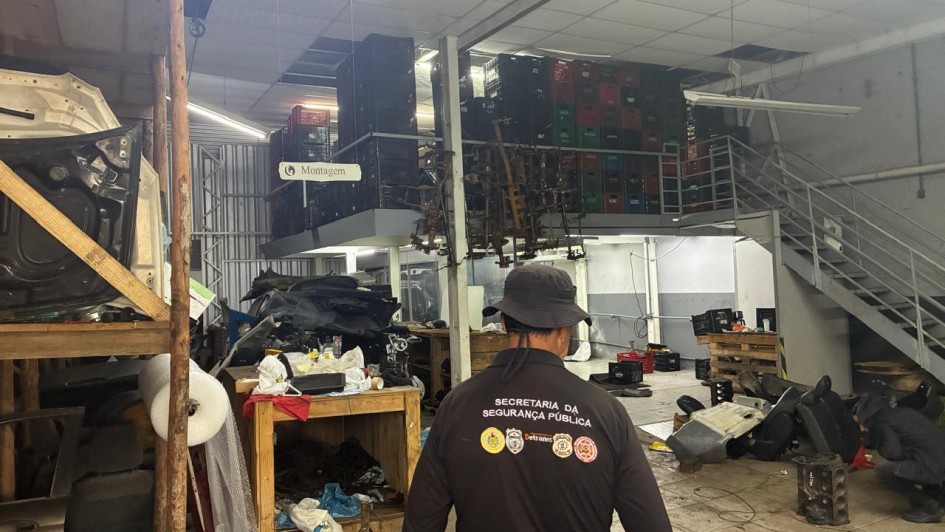 Uma fotografia colorida em plano geral e &acirc;ngulo normal mostra o interior de um grande galp&atilde;o ou ferro-velho de autope&ccedil;as durante uma opera&ccedil;&atilde;o de fiscaliza&ccedil;&atilde;o.

O Agente em Primeiro Plano:

No centro da imagem, em primeiro plano e de costas para a c&acirc;mera, destaca-se um agente policial ou de fiscaliza&ccedil;&atilde;o.

Ele veste um uniforme t&aacute;tico composto por blusa preta de manga longa com a inscri&ccedil;&atilde;o em letras mai&uacute;sculas brancas nas costas: "SECRETARIA DA SEGURAN&Ccedil;A P&Uacute;BLICA".

Abaixo do texto, h&aacute; uma fileira de quatro bras&otilde;es e distintivos coloridos institucionais (incluindo os s&iacute;mbolos do Estado do RS, Pol&iacute;cia Civil, DetranRS e Instituto-Geral de Per&iacute;cias).

Ele usa um chap&eacute;u t&aacute;tico do tipo bucket/saf&aacute;ri preto e cinto com coldre e equipamentos operacionais na cintura.

O Interior do Estabelecimento:

O espa&ccedil;o &eacute; amplo e possui p&eacute;-direito alto, piso de concreto/cer&acirc;mica desgastada e paredes de blocos com divis&oacute;rias de chapas met&aacute;licas.

&Agrave; esquerda, h&aacute; estantes de madeira r&uacute;stica e bancadas abarrotadas de pe&ccedil;as de ve&iacute;culos, como portas de carros, pain&eacute;is, blocos de motor, estofamentos de bancos e rolos de pl&aacute;stico-bolha. Uma placa branca suspensa traz a indica&ccedil;&atilde;o "Montagem" com uma seta apontada para a esquerda.

Ao fundo e no mezanino met&aacute;lico superior, h&aacute; centenas de caixas pl&aacute;sticas organizadoras (pretas, verdes e vermelhas) empilhadas at&eacute; o teto, al&eacute;m de ferragens e eixos veiculares suspensos.

&Agrave; direita, uma escada met&aacute;lica aberta d&aacute; acesso ao mezanino superior. No piso t&eacute;rreo ao lado da escada, outro agente ou operador agacha-se pr&oacute;ximo a bancos automotivos e blocos de motor espalhados pelo ch&atilde;o para inspecionar as pe&ccedil;as. A ilumina&ccedil;&atilde;o &eacute; fornecida por lumin&aacute;rias tubulares fluorescentes e l&acirc;mpadas de LED no teto de forro modular.