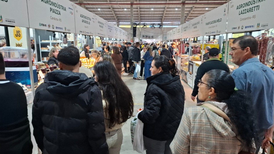 Foto geral de um pavilh&atilde;o, com v&aacute;rias pessoas de p&eacute; pelos corredores olhando os produtos expostos nas bancas.