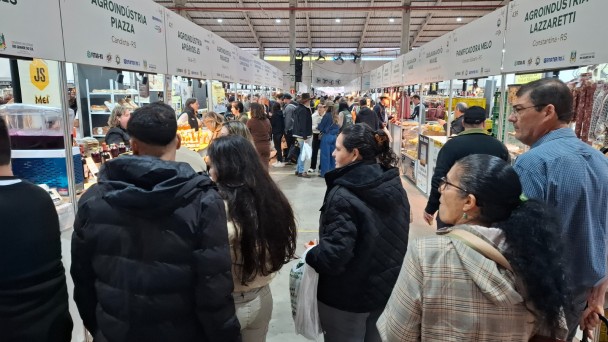 Foto geral de um pavilh&atilde;o, com v&aacute;rias pessoas de p&eacute; pelos corredores olhando os produtos expostos nas bancas.