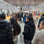 Foto geral de um pavilh&atilde;o, com v&aacute;rias pessoas de p&eacute; pelos corredores olhando os produtos expostos nas bancas.
