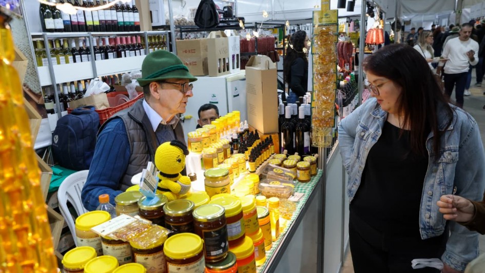Uma fotografia colorida em plano m&eacute;dio foca na intera&ccedil;&atilde;o entre um produtor e uma cliente em um estande do Pavilh&atilde;o da Agricultura Familiar.

**O Produtor e o Balc&atilde;o (&agrave; esquerda e ao centro):**

* &Agrave; esquerda, atr&aacute;s do balc&atilde;o de vendas, um senhor de pele clara e &oacute;culos de grau atende a cliente. Ele usa um chap&eacute;u tradicional de feltro verde com cord&atilde;o escuro, camisa social clara sob um su&eacute;ter de tric&ocirc; azul e colete acolchoado cinza.
* Sobre o balc&atilde;o, h&aacute; uma farta exposi&ccedil;&atilde;o de produtos da apicultura: dezenas de potes de vidro com mel em tons dourados e &acirc;mbar, fechados com tampas amarelas em diferentes tamanhos, al&eacute;m de pequenos frascos conta-gotas de pr&oacute;polis e embalagens com favos de mel.
* Entre os potes, destaca-se um boneco de croch&ecirc; (*amigurumi*) em formato de abelha amarela e preta.
* Ao fundo do estande, veem-se estantes brancas repletas de garrafas de vinho, caixas de papel&atilde;o kraft e outro homem trabalhando ao lado. Fios com luzes decorativas amarelas iluminam o espa&ccedil;o.

**A Cliente e o Corredor (&agrave; direita):**

* &Agrave; direita, em p&eacute; no corredor em frente ao balc&atilde;o, uma mulher de cabelos castanhos longos e &oacute;culos de arma&ccedil;&atilde;o fina observa os produtos com aten&ccedil;&atilde;o. Ela veste uma jaqueta jeans clara sobre blusa preta e cal&ccedil;a escura, segurando uma sacola pl&aacute;stica na m&atilde;o esquerda.
* Pr&oacute;ximo a ela, no canto inferior direito, v&ecirc;-se a m&atilde;o de outra pessoa segurando uma pequena colher de degusta&ccedil;&atilde;o.
* Mais ao fundo, no corredor movimentado do pavilh&atilde;o, outras pessoas e visitantes circulam entre os estandes sob uma lona clara de cobertura de evento.