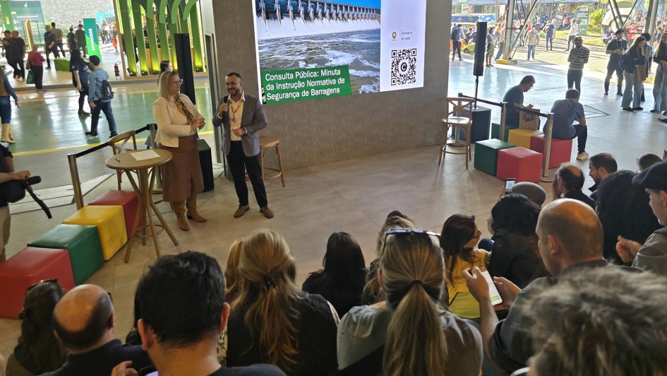 Foto mostra um homem ao microfone ao lado de uma mulher, eles falam para uma plateia em um ambiente coberto mas aberto para a &aacute;rea externa. Ao fundo, um tel&atilde;o com informa&ccedil;&otilde;es sobre a consulta p&uacute;blica.