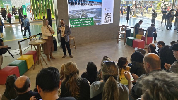 Foto mostra um homem ao microfone ao lado de uma mulher, eles falam para uma plateia em um ambiente coberto mas aberto para a &aacute;rea externa. Ao fundo, um tel&atilde;o com informa&ccedil;&otilde;es sobre a consulta p&uacute;blica.