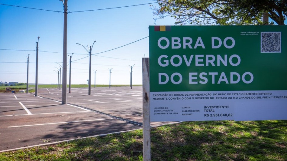 Uma fotografia colorida em plano m&eacute;dio e &acirc;ngulo frontal foca em uma placa oficial de obra p&uacute;blica instalada ao lado de um amplo p&aacute;tio de estacionamento rec&eacute;m-pavimentado.

**A Placa de Obra (em primeiro plano &agrave; direita):**

* No lado direito da imagem, sobressai uma grande placa retangular com topo em tom verde-escuro e base branca.
* No topo verde, l&ecirc;-se em letras garrafais brancas mai&uacute;sculas: **"OBRA DO GOVERNO DO ESTADO"**, com uma pequena bandeira do Rio Grande do Sul no canto superior esquerdo e um QR Code no canto superior direito.
* Na parte inferior branca, constam os detalhes t&eacute;cnicos e institucionais da interven&ccedil;&atilde;o:
* **Descri&ccedil;&atilde;o:** *"EXECU&Ccedil;&Atilde;O DE OBRAS DE PAVIMENTA&Ccedil;&Atilde;O DO P&Aacute;TIO DE ESTACIONAMENTO EXTERNO, MEDIANTE CONV&Ecirc;NIO COM O GOVERNO DO ESTADO DO RIO GRANDE DO SUL FPE N.&ordm; 1330/2025"*.
* **Munic&iacute;pio:** *"SANTO &Acirc;NGELO/RS"*.
* **Empresa Executora:** *"CARPENEDO & CIA LTDA"*.
* **Investimento Total:** *"R$ 2.931.648,82"*.



**O Estacionamento e o Entorno (&agrave; esquerda e ao fundo):**

* &Agrave; esquerda, estende-se o amplo p&aacute;tio de asfalto novo e escuro, marcado por demarca&ccedil;&otilde;es brancas de vagas de estacionamento rec&eacute;m-pintadas.
* Ao longo do p&aacute;tio, alinham-se altos postes de concreto cinza com lumin&aacute;rias de ilumina&ccedil;&atilde;o p&uacute;blica e fia&ccedil;&atilde;o disposta no topo.
* Ao fundo, avista-se uma vegeta&ccedil;&atilde;o rasteira e terreno plano sob um c&eacute;u limpo de tom azul-vivo, sob ilumina&ccedil;&atilde;o solar direta. 
