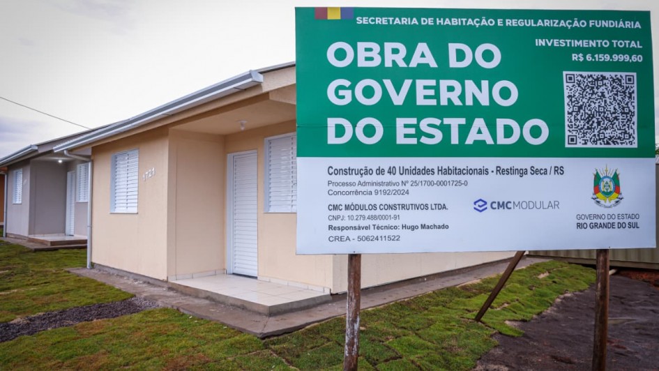 Uma fotografia colorida em plano m&eacute;dio e &acirc;ngulo frontal foca na placa oficial de obra p&uacute;blica instalada em frente a uma resid&ecirc;ncia rec&eacute;m-constru&iacute;da num loteamento habitacional.

**A Placa de Obra (em primeiro plano &agrave; direita):**

* No lado direito da imagem, sobressai uma grande placa retangular com topo em tom verde-escuro e base branca.


* No topo verde, destaca-se no cabe&ccedil;alho o texto **"SECRETARIA DE HABITA&Ccedil;&Atilde;O E REGULARIZA&Ccedil;&Atilde;O FUNDI&Aacute;RIA"** e, abaixo em letras garrafais brancas mai&uacute;sculas: **"OBRA DO GOVERNO DO ESTADO"**. No canto superior direito, l&ecirc;-se **"INVESTIMENTO TOTAL R$ 6.159.999,60"** e um QR Code.


* Na parte inferior branca, constam as informa&ccedil;&otilde;es t&eacute;cnicas e institucionais:


* **T&iacute;tulo:** *"Constru&ccedil;&atilde;o de 40 Unidades Habitacionais - Restinga S&ecirc;ca / RS"*.


* **Processo/Concorr&ecirc;ncia:** *"Processo Administrativo N.&ordm; 25/1700-0001725-0 | Concorr&ecirc;ncia 9192/2024"*.


* **Empresa e Respons&aacute;vel:** *"CMC M&Oacute;DULOS CONSTRUTIVOS LTDA. | Respons&aacute;vel T&eacute;cnico: Hugo Machado CREA - 5062411522"*.


* **Log&oacute;tipos:** A marca *"CMC MODULAR"* ao centro e o bras&atilde;o oficial do *"GOVERNO DO ESTADO RIO GRANDE DO SUL"* no canto inferior direito.





**A Casa e o Entorno (&agrave; esquerda e ao fundo):**

* &Agrave; esquerda, em segundo plano, v&ecirc;-se a fachada de uma casa t&eacute;rrea novinha em folha com paredes pintadas em tom bege-claro/areia e n&uacute;mero residencial **"1721"** fixado na parede.


* O im&oacute;vel possui porta de entrada e janelas com venezianas brancas fechadas, marquise com l&acirc;mpada acesa e calha prateada no beiral do telhado.


* &Agrave; frente da resid&ecirc;ncia, estende-se uma faixa de relva rec&eacute;m-plantada e caminho de acesso em concreto. Ao fundo, avista-se a continua&ccedil;&atilde;o da fileira de casas do conjunto habitacional sob um c&eacute;u nublado.