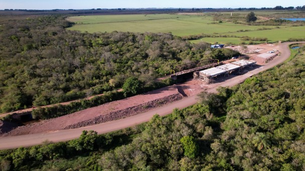 Uma fotografia a&eacute;rea colorida em plano geral e perspetiva obl&iacute;qua (capturada por drone) regista a vista panor&acirc;mica das obras de infraestrutura e reconstru&ccedil;&atilde;o de uma ponte rodovi&aacute;ria num ambiente rural e nativo.

**A Ponte e o Canteiro de Obras (ao centro e &agrave; direita):**

* No centro-direito da composi&ccedil;&atilde;o, observa-se a estrutura em constru&ccedil;&atilde;o de uma nova ponte de bet&atilde;o armado, com os tabuleiros e pilares de sustenta&ccedil;&atilde;o montados.


* Paralelamente &agrave; nova ponte, encontra-se a estrutura met&aacute;lica de uma antiga ponte treli&ccedil;ada de tom escuro.


* Ao lado das estruturas, estende-se uma via de terra batida e cascalho avermelhado. Na extremidade superior da curva, avista-se o canteiro de apoio com contentores, ve&iacute;culos e materiais de constru&ccedil;&atilde;o organizados.



**A Paisagem e a Vegeta&ccedil;&atilde;o:**

* A via e as pontes cruzam uma vasta &aacute;rea rodeada por uma densa mata nativa e &aacute;rvores de copa verde-escura.


* Ao fundo e em redor da &aacute;rea florestal, a paisagem abre-se em extensos campos de cultivo verdes e pastagens sob a linha do horizonte, sob ilumina&ccedil;&atilde;o solar direta e c&eacute;u claro num tom azul-suave.