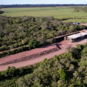 Uma fotografia a&eacute;rea colorida em plano geral e perspetiva obl&iacute;qua (capturada por drone) regista a vista panor&acirc;mica das obras de infraestrutura e reconstru&ccedil;&atilde;o de uma ponte rodovi&aacute;ria num ambiente rural e nativo.

**A Ponte e o Canteiro de Obras (ao centro e &agrave; direita):**

* No centro-direito da composi&ccedil;&atilde;o, observa-se a estrutura em constru&ccedil;&atilde;o de uma nova ponte de bet&atilde;o armado, com os tabuleiros e pilares de sustenta&ccedil;&atilde;o montados.


* Paralelamente &agrave; nova ponte, encontra-se a estrutura met&aacute;lica de uma antiga ponte treli&ccedil;ada de tom escuro.


* Ao lado das estruturas, estende-se uma via de terra batida e cascalho avermelhado. Na extremidade superior da curva, avista-se o canteiro de apoio com contentores, ve&iacute;culos e materiais de constru&ccedil;&atilde;o organizados.



**A Paisagem e a Vegeta&ccedil;&atilde;o:**

* A via e as pontes cruzam uma vasta &aacute;rea rodeada por uma densa mata nativa e &aacute;rvores de copa verde-escura.


* Ao fundo e em redor da &aacute;rea florestal, a paisagem abre-se em extensos campos de cultivo verdes e pastagens sob a linha do horizonte, sob ilumina&ccedil;&atilde;o solar direta e c&eacute;u claro num tom azul-suave.