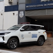 Uma fotografia colorida em plano m&eacute;dio e perspectiva em tr&ecirc;s quartos capta uma viatura oficial de Per&iacute;cia Criminal estacionada em frente &agrave; sede do Instituto-Geral de Per&iacute;cias do Rio Grande do Sul (IGP-RS).

**A Viatura Oficial (em primeiro plano):**

* No centro e &agrave; esquerda da imagem, destaca-se um ve&iacute;culo utilit&aacute;rio desportivo (SUV) oficial da marca Toyota, de cor branca, visto de perfil diagonal de tr&ecirc;s quartos.
* Na porta lateral do condutor, v&ecirc;-se o bras&atilde;o do **"INSTITUTO-GERAL DE PER&Iacute;CIAS RS"** e a inscri&ccedil;&atilde;o em letras azuis e pretas: **"PER&Iacute;CIA CRIMINAL - RIO GRANDE DO SUL"**.
* Junto &agrave; janela traseira, observa-se a bandeira do Estado do Rio Grande do Sul. No teto do ve&iacute;culo, encontra-se instalado um sinalizador luminoso (giroflex) de emerg&ecirc;ncia em tom vermelho-escuro.

 O Edif&iacute;cio do IGP (ao fundo e &agrave; direita):

* Ao fundo, ergue-se a fachada do pr&eacute;dio oficial. Na parte superior da entrada, sobre uma faixa de azulejos ou pastilhas azuis, sobressai a inscri&ccedil;&atilde;o em letras garrafais brancas mai&uacute;sculas: **"INSTITUTO-GERAL DE PER&Iacute;CIAS DO RI..."**
* Num pilar cinzento &agrave; esquerda da entrada principal, est&aacute; fixado o bras&atilde;o oficial do Instituto-Geral de Per&iacute;cias.
* &Agrave; direita, avista-se a entrada do edif&iacute;cio com portas de vidro, escadaria de acesso com corrim&atilde;o met&aacute;lico prateado e rampas. O piso exterior &eacute; feito de lajotas de pedra sob ilumina&ccedil;&atilde;o natural diurna.