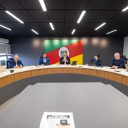 Uma fotografia colorida em plano geral e perspetiva em &acirc;ngulo levemente baixo capta o governador Eduardo Leite reunido com secret&aacute;rios de Estado ao redor de uma grande mesa semicircular de reuni&otilde;es numa sala institucional.

**A Mesa Principal e as Autoridades (ao centro e em primeiro plano):**

* Ao centro da mesa semicircular em tom de madeira clara, o governador Eduardo Leite fala ao microfone com as m&atilde;os entrela&ccedil;adas. Ele veste blazer escuro sobre camisa clara.


* Ao redor da mesa, dispostos em curva, secret&aacute;rios e secret&aacute;rias acompanham a reuni&atilde;o com computadores port&aacute;teis, tablets e blocos de anota&ccedil;&otilde;es sobre a bancada, al&eacute;m de identificadores nominais &agrave; frente de cada lugar.


* &Agrave; esquerda do governador, v&ecirc;-se um homem de blazer azul e, na extremidade esquerda, uma mulher de casaco de ganga a trabalhar num port&aacute;til.


* &Agrave; direita do governador, encontram-se outros quatro secret&aacute;rios e assessores em trajes formais e esporte fino (blazers e camisas sociais), observando a apresenta&ccedil;&atilde;o ou consultando os seus dispositivos eletr&oacute;nicos.




* Em primeiro plano, no centro da composi&ccedil;&atilde;o, sobressai o tampo cinzento de um m&oacute;dulo da mesa com uma folha impressa onde se l&ecirc; **"SALA RIO GRANDE"**.



**O Ambiente e a S&iacute;mbolo Oficial (ao fundo):**

* Na parede do fundo, em posi&ccedil;&atilde;o central de destaque atr&aacute;s do governador, sobressai um grande painel retangular com a bandeira do Estado do Rio Grande do Sul e o seu bras&atilde;o de armas impresso no centro.


* A sala possui paredes escuras e teto preto com linhas paralelas de ilumina&ccedil;&atilde;o LED encastradas e acesas, garantindo uma luz clara e focada sobre a mesa de reuni&otilde;es. &Agrave; esquerda, avistam-se janelas com persianas cinzentas.