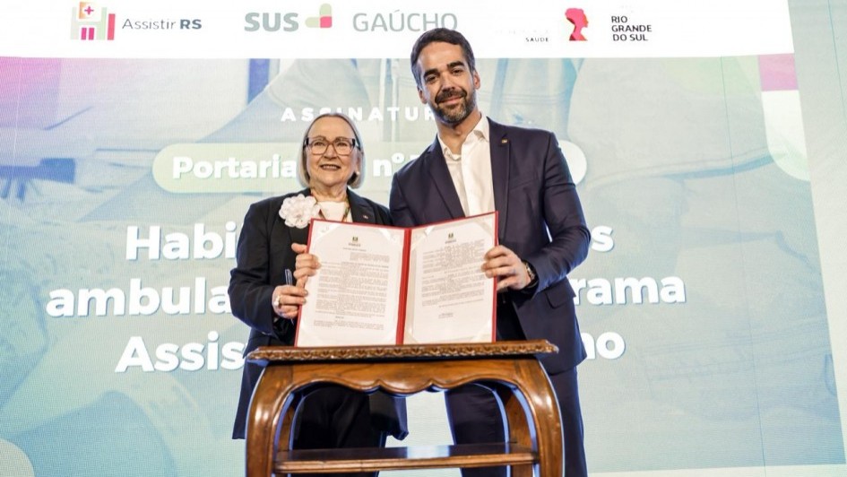 Uma fotografia colorida em plano m&eacute;dio e perspetiva ao n&iacute;vel dos olhos regista o momento de uma assinatura solene de portaria pelo governador Eduardo Leite e pela secret&aacute;ria da Sa&uacute;de, Arita Bergmann.

**As Autoridades e o Documento (ao centro):**

* Ao centro, o governador Eduardo Leite (&agrave; direita) e a secret&aacute;ria estadual da Sa&uacute;de, Arita Bergmann (&agrave; esquerda), posam sorridentes segurando juntos uma pasta vermelha aberta contendo o documento oficial assinado.
* O governador veste fato azul-marinho, camisa branca sem gravata e pin institucional na lapela.
* A secret&aacute;ria veste blazer azul-escuro com uma flor branca decorativa na lapela, blusa clara, &oacute;culos de arma&ccedil;&atilde;o preta e acess&oacute;rios prateados.
* &Agrave; frente do par, encontra-se uma pequena mesa de madeira nobre em tom castanho-m&eacute;dio com pernas curvadas de estilo cl&aacute;ssico.

**O Painel de Fundo e as Marcas Institucionais (ao fundo):**

* Ao fundo, destaca-se um grande ecran LED com proje&ccedil;&atilde;o em tons claros e azuis com o t&iacute;tulo do evento: **"ASSINATURA"**, seguido do texto em destaque **"Portaria n&ordm;..."** e trechos como **"Habilita&ccedil;&atilde;o de servi&ccedil;os ambulat&oacute;rias... Programa Assistir"**.
* Na parte superior do painel, exibem-se os log&oacute;tipos do programa **"Assistir RS"**, **"SUS GA&Uacute;CHO"**, da **"SECRETARIA DA SA&Uacute;DE"** e do Governo do Estado do **"RIO GRANDE DO SUL"**