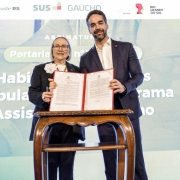 Uma fotografia colorida em plano m&eacute;dio e perspetiva ao n&iacute;vel dos olhos regista o momento de uma assinatura solene de portaria pelo governador Eduardo Leite e pela secret&aacute;ria da Sa&uacute;de, Arita Bergmann.

**As Autoridades e o Documento (ao centro):**

* Ao centro, o governador Eduardo Leite (&agrave; direita) e a secret&aacute;ria estadual da Sa&uacute;de, Arita Bergmann (&agrave; esquerda), posam sorridentes segurando juntos uma pasta vermelha aberta contendo o documento oficial assinado.
* O governador veste fato azul-marinho, camisa branca sem gravata e pin institucional na lapela.
* A secret&aacute;ria veste blazer azul-escuro com uma flor branca decorativa na lapela, blusa clara, &oacute;culos de arma&ccedil;&atilde;o preta e acess&oacute;rios prateados.
* &Agrave; frente do par, encontra-se uma pequena mesa de madeira nobre em tom castanho-m&eacute;dio com pernas curvadas de estilo cl&aacute;ssico.

**O Painel de Fundo e as Marcas Institucionais (ao fundo):**

* Ao fundo, destaca-se um grande ecran LED com proje&ccedil;&atilde;o em tons claros e azuis com o t&iacute;tulo do evento: **"ASSINATURA"**, seguido do texto em destaque **"Portaria n&ordm;..."** e trechos como **"Habilita&ccedil;&atilde;o de servi&ccedil;os ambulat&oacute;rias... Programa Assistir"**.
* Na parte superior do painel, exibem-se os log&oacute;tipos do programa **"Assistir RS"**, **"SUS GA&Uacute;CHO"**, da **"SECRETARIA DA SA&Uacute;DE"** e do Governo do Estado do **"RIO GRANDE DO SUL"**