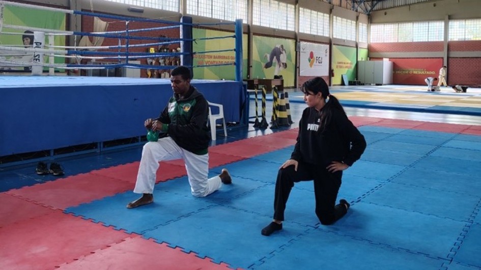 Uma fotografia colorida em plano geral e perspectiva ao n&iacute;vel dos olhos capta uma sess&atilde;o de fisioterapia esportiva e treino funcional nas instala&ccedil;&otilde;es do Centro Estadual de Treinamento Esportivo (Cete).

**Os Atletas e o Exerc&iacute;cio (ao centro e em primeiro plano):**

* No centro da composi&ccedil;&atilde;o, sobre tatames emborrachados em tons de azul e vermelho, duas pessoas realizam um exerc&iacute;cio de afundo/alongamento ajoelhado:
* &Agrave; esquerda, um homem negro veste casaco desportivo verde e preto, cal&ccedil;as brancas e segura um *kettlebell* verde com a m&atilde;o direita enquanto mant&eacute;m o joelho esquerdo fletido &agrave; frente.
* &Agrave; direita, uma jovem de cabelos escuros apanhados num rabo de cavalo veste camisola de manga comprida preta com a marca **"PUMA"**, cal&ccedil;as desportivas pretas e meia-p&eacute;s escuros, apoiando a m&atilde;o no joelho fletido.

**A Infraestrutura do Gin&aacute;sio (&agrave; esquerda e ao fundo):**

* &Agrave; esquerda, em segundo plano, destaca-se um ringue de boxe elevado com lona azul e cordas azuis e brancas.
* Ao fundo e &agrave; direita, observa-se a ampla &aacute;rea de treino com tatames e &aacute;reas destinadas a artes marciais (jud&ocirc;/m&uacute;ltiplas modalidades), com banners institucionais fixados nas paredes e o log&oacute;tipo da **FGJ** (Federa&ccedil;&atilde;o Ga&uacute;cha de Jud&ocirc;).
* O pavilh&atilde;o conta com p&eacute;-direito alto, estrutura met&aacute;lica no teto com tesouras em treli&ccedil;a, janelas altas com ilumina&ccedil;&atilde;o natural e tijolos &agrave; vista na parte inferior das paredes.
