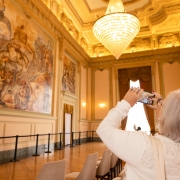 Uma fotografia colorida em plano m&eacute;dio-geral com perspetiva em contra-plong&eacute;e (de baixo para cima) capta uma visitante a fotografar o interior suntuoso do Pal&aacute;cio Piratini durante uma visita guiada.

**A Visitante (em primeiro plano e &agrave; direita):**

* Em primeiro plano, do lado direito, v&ecirc;-se uma mulher de costas e perfil com cabelos curtos grisalhos/platinados, vestindo uma camisola branca com detalhes em renda nos ombros.


* Ela segura um smartphone na horizontal com ambas as m&atilde;os, elevando o dispositivo para fotografar o teto e os afrescos do sal&atilde;o.



**A Arquitetura e os Afrescos (&agrave; esquerda e ao fundo):**

* &Agrave; esquerda, em grande destaque na parede lateral, exibe-se um enorme afresco hist&oacute;rico em tons cl&aacute;ssicos (obra do pintor Aldo Locatelli), retratando figuras hist&oacute;ricas e cenas da forma&ccedil;&atilde;o do Estado.


* No canto inferior esquerdo, um homem de t-shirt branca e cal&ccedil;as escuras contempla a pintura atr&aacute;s de uma fita de isolamento pedestre com pedestais pretos.


* O sal&atilde;o nobre possui paredes ornamentadas com molduras douradas, colunas decorativas e janelas altas cobertas por cortinados elegantes em tom castanho-dourado.


* O piso &eacute; em taco de madeira polida disposta em parquet.



**A Ilumina&ccedil;&atilde;o e o Teto:**

* Pendente do teto trabalhado em gesso com relevos e detalhes dourados, sobressai um imponente candelabro de cristal aceso, projetando uma luz quente, dourada e acolhedora sobre todo o espa&ccedil;o.
