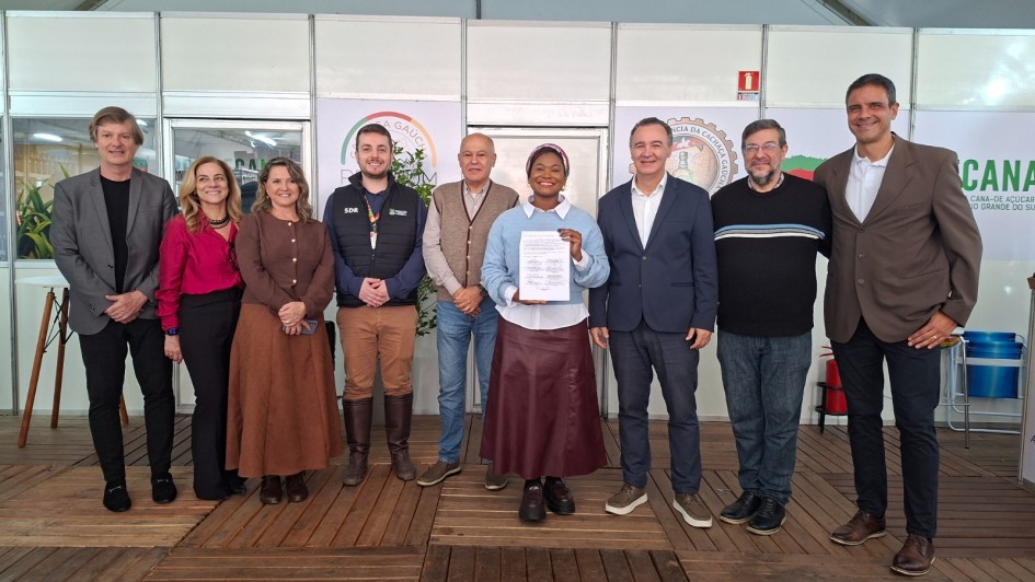 Uma fotografia colorida em plano geral e perspetiva ao n&iacute;vel dos olhos capta um grupo de autoridades e representantes reunidos num estande institucional durante um evento oficial ou feira agr&iacute;cola.

**O Grupo e a Apresenta&ccedil;&atilde;o do Documento (ao centro):**

* No centro da composi&ccedil;&atilde;o, sobressai uma mulher negra de fato/turbante colorido na cabe&ccedil;a, vestindo camisola azul-clara sobre camisa branca, saia longa plissada em tom vi&ntilde;o/castanho-escuro e cal&ccedil;ado escuro.


* Ela segura e exibe com ambas as m&atilde;os um documento oficial impresso assinado.


* Ao seu lado e distribu&iacute;dos alinhados em fila horizontal, posam sorridentes outros sete homens e duas mulheres em trajes formais e esporte fino (blazers, casacos, camisas sociais e coletes):


* &Agrave; esquerda, veem-se duas mulheres (uma com camisa cor-de-rosa vibrante e cal&ccedil;as escuras, e outra com vestido/conjunto castanho) ao lado de integrantes do governo e do setor rural (um deles vestindo colete com a sigla **"SDR"** e botas de couro).


* &Agrave; direita, alinham-se gestores e autoridades vestindo blazers em tom azul-marinho, camisolas escuras com listras brancas e um homem &agrave; extrema-direita de blazer castanho-claro e cal&ccedil;as escuras.





 O Cen&aacute;rio e a Estrutura do Estande (ao fundo):

* O grupo encontra-se sobre um estrado/piso de t&aacute;buas de madeira num estande de exposi&ccedil;&atilde;o.


* Ao fundo, a parede do estande &eacute; formada por pain&eacute;is brancos modulares com divis&oacute;rias met&aacute;licas.


* Sobre os pain&eacute;is, observam-se selos e log&oacute;tipos circulares institucionais, incluindo refer&ecirc;ncias a produtos e cadeias produtivas locais (como a cacha&ccedil;a ga&uacute;cha e a cana-de-a&ccedil;&uacute;car).


* &Agrave; direita, ao fundo, sobressai um banner verde com a inscri&ccedil;&atilde;o parcial **"CANA"** e **"CANA-DE-A&Ccedil;&Uacute;CAR NO RIO GRANDE DO SUL"**. A ilumina&ccedil;&atilde;o natural &eacute; clara e uniforme.