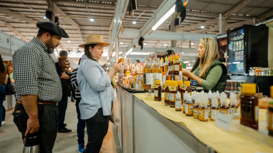 A imagem mostra um estande em um pavilh&atilde;o com produtos da agricultura familiar. Um casal observa os produtos e uma vendedora est&aacute; posicionada atr&aacute;s do balc&atilde;o. 