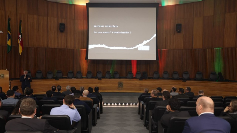 Foto de um audit&oacute;rio com diversas pessoas sentadas em poltronas e um grande painel com a apresenta&ccedil;&atilde;o ao centro do palco. 