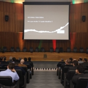Foto de um audit&oacute;rio com diversas pessoas sentadas em poltronas e um grande painel com a apresenta&ccedil;&atilde;o ao centro do palco. 