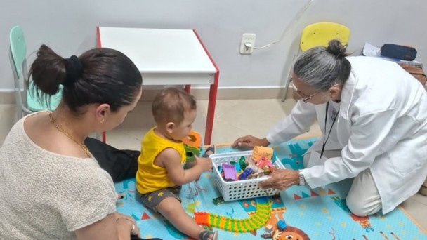 Uma fotografia colorida em plano m&eacute;dio e perspectiva em &acirc;ngulo levemente elevado regista uma sess&atilde;o de atendimento especializado infantil do programa AcompanhaRAPS em Iju&iacute;.

**Os Participantes e a A&ccedil;&atilde;o (em primeiro e meio plano):**

* No centro da composi&ccedil;&atilde;o, sobre um tapete infantil ilustrado e colorido em tons azuis com desenhos de animais e trilhos, tr&ecirc;s pessoas est&atilde;o sentadas ao n&iacute;vel do ch&atilde;o:


* &Agrave; esquerda, em primeiro plano, uma mulher de costas e perfil de cabelos escuros apanhados num coque veste camisola bege de malha texturizada e acompanha o filho.


* No centro, um beb&eacute; de camisa amarela sem mangas e cal&ccedil;&otilde;es escuros brinca no ch&atilde;o com blocos pedag&oacute;gicos de encaixe e brinquedos coloridos.


* &Agrave; direita, uma profissional de sa&uacute;de de cabelos grisalhos apanhados, vestindo bata/jaleco branco sobre roupa clara e crach&aacute; ao pesco&ccedil;o, est&aacute; ajoelhada e aproxima com cuidado um cesto pl&aacute;stico branco repleto de brinquedos pedag&oacute;gicos para a crian&ccedil;a.

**O Ambiente e o Mobili&aacute;rio (ao fundo):**

* Ao fundo, v&ecirc;-se uma pequena mesa infantil quadrada com tampo branco e pernas vermelhas.

* Ao lado da mesa, observam-se duas cadeiras infantis em tons de verde-menta e amarelo.

* As paredes da sala de atendimento s&atilde;o claras e lisas, com tomada el&eacute;trica vis&iacute;vel e ilumina&ccedil;&atilde;o interior direta e acolhedora.