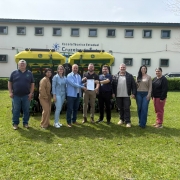 Foto mostra autoridades reunidas e segurando o documento em frente &agrave; escola. Eles est&atilde;o em um gramado verde. A escola tem fachada branca. 