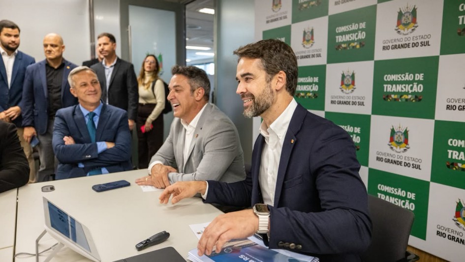 Reuni&atilde;o em ambiente institucional, com participantes sentados &agrave; mesa diante de um painel com os logotipos do Governo do Estado e da Comiss&atilde;o de Transi&ccedil;&atilde;o. Sobre a mesa h&aacute; documentos, dispositivos eletr&ocirc;nicos e materiais de trabalho utilizados durante o encontro.