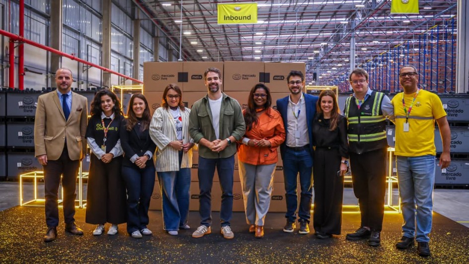 Uma fotografia colorida em plano geral e perspetiva ao n&iacute;vel dos olhos capta a inaugura&ccedil;&atilde;o do centro de distribui&ccedil;&atilde;o do Mercado Livre em Gua&iacute;ba, com a presen&ccedil;a do governador Eduardo Leite, executivos e autoridades.

**As Autoridades e Executivos (ao centro e em primeiro plano):**

* No centro de um palco elevado coberto de alcatifa escura com confetis dourados espalhados pelo ch&atilde;o, dez pessoas posam alinhadas lado a lado para a fotografia oficial.


* Ao centro, o governador Eduardo Leite sorri com as m&atilde;os entrela&ccedil;adas &agrave; frente, vestindo sobrecamisa verde-oliveira sobre t-shirt branca, cal&ccedil;as de ganga escuras e sapatilhas claras.


* &Agrave; sua esquerda (&agrave; direita do observador), encontra-se uma mulher de casaco/blusa de tom laranja-terracota e cal&ccedil;as cinzentas, seguida por executivos de blazer e trajes formais.


* Na extremidade direita, um integrante da equipa veste a t-shirt amarela oficial do Mercado Livre com fita refletora e crach&aacute;, ao lado de um gestor com colete de seguran&ccedil;a refletor de alta visibilidade.


* &Agrave; direita de Eduardo Leite (&agrave; esquerda do observador), alinham-se quatro mulheres em trajes esporte fino e blazer (incluindo uma com t-shirt preta onde se l&ecirc; **"Oi, cheguei! :-)"**) e um homem no extremo esquerdo de blazer bege e gravata.



**O Cen&aacute;rio do Centro de Distribui&ccedil;&atilde;o (ao fundo):**

* Diretamente atr&aacute;s do grupo, ergue-se uma pilha sim&eacute;trica de caixas de cart&atilde;o do **"mercado livre"** e **"meli+"**, emoldurada por estruturas c&uacute;bicas decorativas de ilumina&ccedil;&atilde;o LED amarela/dourada.


* Acima do palco, pendem placas sinalizadoras amarelas de setores do armaz&eacute;m com a palavra **"Inbound"** e frases institucionais.


* Ao fundo e em redor, estende-se a vasta infraestrutura do centro log&iacute;stico com p&eacute;-direito alto, estrutura met&aacute;lica no teto, tubagens industriais e extensas fileiras de estantes/porta-paletes azuis e laranjas.