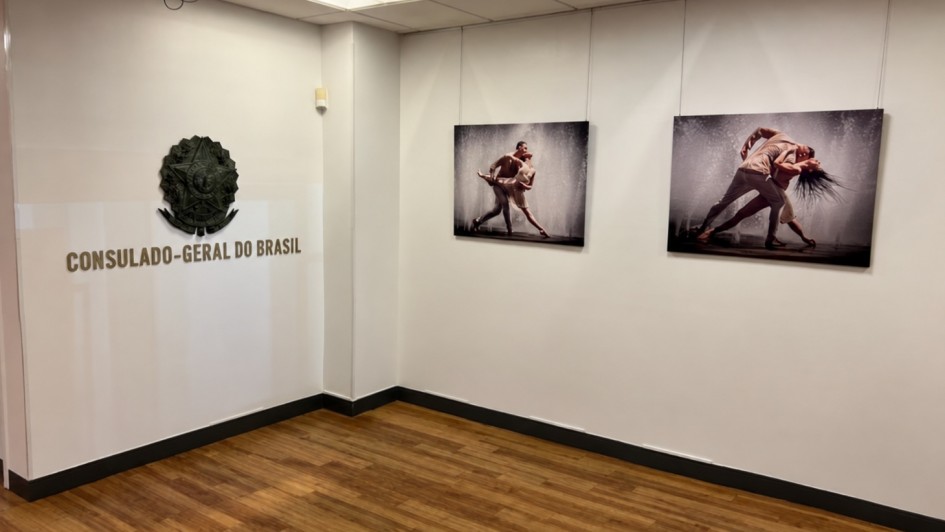 Uma fotografia colorida em plano geral e perspectiva em &acirc;ngulo levemente elevado capta a &aacute;rea de exposi&ccedil;&otilde;es no Consulado-Geral do Brasil em Nova Iorque, sem a presen&ccedil;a de pessoas.

**A Identifica&ccedil;&atilde;o Institucional (&agrave; esquerda):**

* Na parede branca do lado esquerdo, destaca-se em relevo met&aacute;lico escuro o Bras&atilde;o da Rep&uacute;blica Federativa do Brasil.


* Logo abaixo do bras&atilde;o, l&ecirc;-se a inscri&ccedil;&atilde;o em letras garrafais mai&uacute;sculas met&aacute;licas: **"CONSULADO-GERAL DO BRASIL"**.



**A Galeria e as Obras Expostas (ao fundo):**

* Na parede branca do fundo, encontram-se expostas duas fotografias art&iacute;sticas retangulares de grande formato, suspensas por finos cabos met&aacute;licos presos ao teto:


* A fotografia da esquerda retrata um casal de bailarinos numa pose de dan&ccedil;a expressiva sob jorros de &aacute;gua/chuva iluminados.


* A fotografia da direita exibe o mesmo casal numa pose din&acirc;mica de dan&ccedil;a contempor&acirc;nea, com movimentos dram&aacute;ticos e salpicos de &aacute;gua ao fundo.





**O Espa&ccedil;o e a Ilumina&ccedil;&atilde;o:**

* O espa&ccedil;o possui piso em reguado de madeira em tom castanho-m&eacute;dio com acabamento acetinado e rodap&eacute;s escuros ao longo das paredes brancas e lisas.


* O teto modular em placas brancas conta com pain&eacute;is de ilumina&ccedil;&atilde;o encastrados que emitem uma luz suave e uniforme sobre a &aacute;rea de exposi&ccedil;&atilde;o.