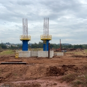 Uma fotografia colorida em plano geral e perspectiva ao n&iacute;vel dos olhos capta o canteiro de obras de constru&ccedil;&atilde;o de uma nova ponte, destacando a etapa de execu&ccedil;&atilde;o dos pilares de sustenta&ccedil;&atilde;o.

**A Infraestrutura em Constru&ccedil;&atilde;o (ao centro):**

* No centro da composi&ccedil;&atilde;o, sobressaem-se dois grandes pilares de sustenta&ccedil;&atilde;o em fase de betonagem e arma&ccedil;&atilde;o de a&ccedil;o, erguidos sobre blocos de funda&ccedil;&atilde;o maci&ccedil;os em bet&atilde;o cinzento-claro.


* A base redonda dos pilares est&aacute; envolvida por cofragens met&aacute;licas azuis, sobre as quais se encontram instaladas plataformas de trabalho e guarda-corpos de prote&ccedil;&atilde;o pintados de amarelo vibrante.


* No topo dos pilares, elevam-se as armaduras de ferro/a&ccedil;o expostas em formato cil&iacute;ndrico, aguardando as pr&oacute;ximas etapas de betonagem.



**O Canteiro de Obras e o Entorno (em primeiro plano e ao fundo):**

* Em primeiro plano e ao redor das funda&ccedil;&otilde;es, estende-se uma ampla &aacute;rea de terra batida e revolvida em tom terroso/castanho-avermelhado com marcas de pneus de m&aacute;quinas pesadas.


* Ao fundo, &agrave; direita, avistam-se m&aacute;quinas escavadoras, perfuradoras e equipamentos de constru&ccedil;&atilde;o civil em atividade no terreno.


* Ao fundo e &agrave; esquerda, observa-se uma &aacute;rea verde com vegeta&ccedil;&atilde;o densa, &aacute;rvores e ao longe a silhueta de edifica&ccedil;&otilde;es e de uma igreja rural.


* O c&eacute;u apresenta-se completamente encoberto por nuvens cinzentas e ilumina&ccedil;&atilde;o natural difusa de dia nublado.
