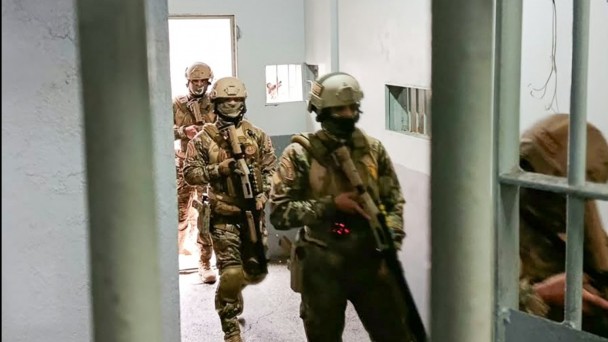 Foto mostra policiais penais com fardas, capacete e m&aacute;scara, usando armas longas, est&atilde;o no corredor de uma unidade prisional.
