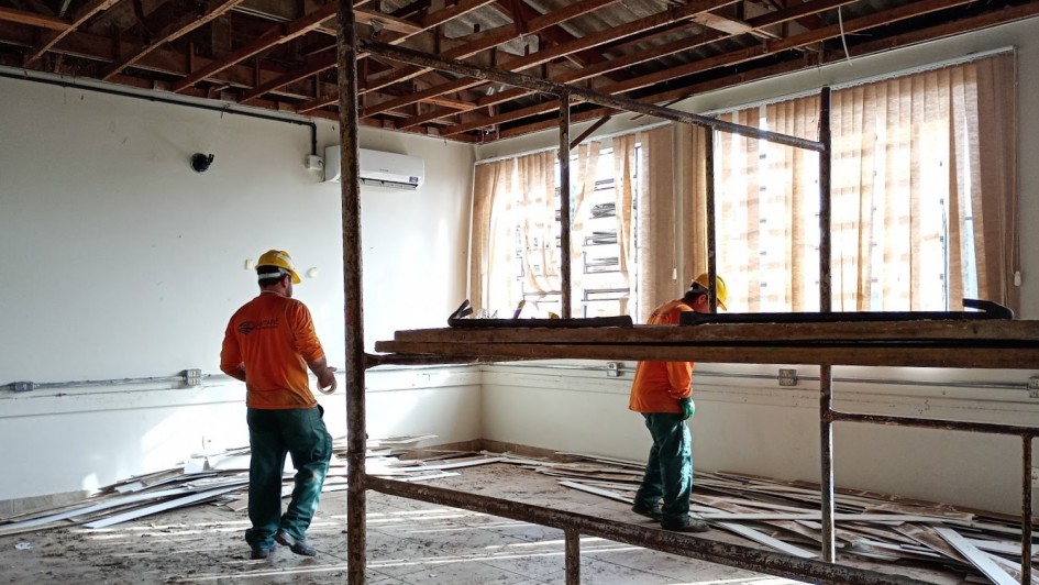 Uma fotografia colorida em plano geral e perspectiva ao n&iacute;vel dos olhos capta trabalhadores da constru&ccedil;&atilde;o civil durante as obras de reforma e recupera&ccedil;&atilde;o de uma sala na Escola Estadual Dem&eacute;trio Ribeiro, em Alegrete.

**Os Trabalhadores e a A&ccedil;&atilde;o (em primeiro e meio plano):**

* No centro e &agrave; esquerda da composi&ccedil;&atilde;o, dois oper&aacute;rios trabalham na desmontagem e remodela&ccedil;&atilde;o do espa&ccedil;o, vestindo camisas de trabalho de manga comprida de cor laranja-vibrante, cal&ccedil;as de prote&ccedil;&atilde;o verdes, capacetes de seguran&ccedil;a amarelos e botas de trabalho.


* O oper&aacute;rio da esquerda caminha de costas em dire&ccedil;&atilde;o &agrave; parede, segurando um rolo de fita adesiva/medida na m&atilde;o direita.


* O oper&aacute;rio da direita est&aacute; posicionado sobre a plataforma de t&aacute;buas de madeira de um andaime met&aacute;lico, trabalhando na remo&ccedil;&atilde;o de materiais do teto.


* Em primeiro plano, destaca-se a estrutura tubular met&aacute;lica em tom castanho/enferrujado de um andaime de dois n&iacute;veis.



**A Estrutura e o Ambiente da Sala:**

* No teto, exp&otilde;e-se a estrutura de ripas de madeira do telhado ap&oacute;s a remo&ccedil;&atilde;o do forro original, com telhas onduladas vis&iacute;veis no topo.


* As paredes est&atilde;o pintadas de branco/creme, com tubagens el&eacute;tricas externas de alum&iacute;nio e tomadas instaladas a meia altura. Na parede do fundo, sobressai um aparelho de ar condicionado do tipo *split* branco.


* Ao fundo, grandes janelas est&atilde;o cobertas por persianas verticais de tecido bege/castanho-claro, filtrando a luz solar intensa do exterior.


* O ch&atilde;o de cer&acirc;mica clara est&aacute; repleto de restos de madeira, ripas e entulho resultantes da desmontagem da cobertura.