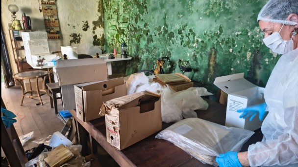 Foto mostra uma t&eacute;cnica de avental, touca e m&aacute;scaras brancas e luvas azuis mexendo num saco com documentos. Na frente dela, h&aacute; v&aacute;rias caixas com documentos. 
