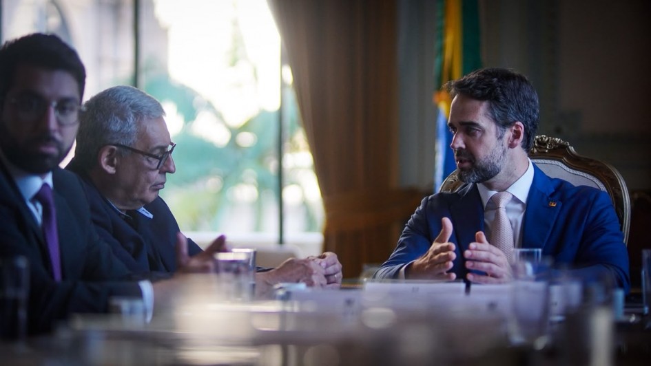Uma fotografia colorida em plano m&eacute;dio-fechado e perspectiva ao n&iacute;vel dos olhos capta o governador Eduardo Leite em reuni&atilde;o com o prefeito de Porto Alegre, Sebasti&atilde;o Melo, no Pal&aacute;cio Piratini.

**O Governador e o Prefeito (ao centro e em primeiro plano):**

* &Agrave; direita, o governador Eduardo Leite fala de perfil diagonal voltado para a esquerda, gesticulando com ambas as m&atilde;os abertas sobre a mesa. Ele veste um fato azul-vibrante, camisa branca, gravata bege/rosa-claro e traz um pequeno pin com a bandeira do Estado na lapela do casaco.
* &Agrave; esquerda, em primeiro plano, o prefeito de Porto Alegre, Sebasti&atilde;o Melo, escuta atentamente de perfil, ligeiramente inclinado para a frente com as m&atilde;os entrela&ccedil;adas sobre a mesa. Ele possui cabelos grisalhos e usa &oacute;culos de arma&ccedil;&atilde;o escura.
* Ao seu lado, no extremo esquerdo em primeiro plano, avista-se de perfil outro gestor de fato escuro, camisa branca e gravata roxa/vinho.
* Sobre a mesa de madeira nobre em primeiro plano, veem-se copos de &aacute;gua e identificadores nominais em acr&iacute;lico desfocados pela profundidade de campo reduzida.

**O Ambiente do Sal&atilde;o (ao fundo):**

* Ao fundo, entre as figuras principais, uma grande janela iluminada revela a vegeta&ccedil;&atilde;o verde do exterior atrav&eacute;s de um vidro amplo e cortinados dourados recolhidos.
* &Agrave; direita de Eduardo Leite, avista-se parcialmente a Bandeira do Rio Grande do Sul (nas cores verde, vermelha e amarela) disposta num hasteamento interior.
* A ilumina&ccedil;&atilde;o natural proveniente da janela ao fundo cria um forte contraste de contraluz, destacando os contornos dos participantes na mesa de reuni&atilde;o.