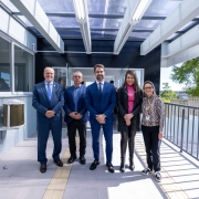 Grupo de pessoas posando em uma passarela coberta de um edif&iacute;cio moderno, com paredes em tons claros, cobertura transl&uacute;cida e guarda-corpo met&aacute;lico; ao fundo, h&aacute; portas envidra&ccedil;adas e vista parcial da &aacute;rea externa.