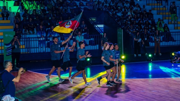 Foto mostra grupo de atletas entrando correndo na quadra. Um deles carrega bem alto a bandeira do Rio Grande do Sul. Ao fundo, na penumbra, aparece a plateia na arquibancada do gin&aacute;sio.