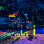 Foto mostra grupo de atletas entrando correndo na quadra. Um deles carrega bem alto a bandeira do Rio Grande do Sul. Ao fundo, na penumbra, aparece a plateia na arquibancada do gin&aacute;sio.