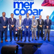 Uma fotografia colorida em plano geral e perspectiva ao n&iacute;vel dos olhos registra o governador Eduardo Leite acompanhado por autoridades e lideran&ccedil;as empresariais no palco de abertura da Mercopar, em Caxias do Sul.

**As Autoridades e Lideran&ccedil;as no Palco (ao centro e em primeiro plano):**

* No centro de um palco coberto por alcatifa azul, sete homens posam alinhados lado a lado para a fotografia oficial com as m&atilde;os entrela&ccedil;adas &agrave; frente do corpo.


* Ao centro, o governador Eduardo Leite sorri de fato azul-bic/azul-vibrante, camisa social branca e gravata azul-marinho.


* &Agrave; sua direita (&agrave; esquerda do observador), alinham-se tr&ecirc;s gestores e lideran&ccedil;as: um homem de fato azul-marinho de &oacute;culos e gravata salm&atilde;o; um gestor de fato azul-escuro sem gravata; e um homem de blazer azul-claro com camisola amarela por baixo de camisa social.


* &Agrave; sua esquerda (&agrave; direita do observador), encontram-se outros tr&ecirc;s representantes: um homem de fato cinzento-claro com gravata verde; um executivo de fato escuro com crach&aacute; oficial do evento; e um homem de fato azul-escuro, camisa clara e gravata escura na extremidade direita.


* Em primeiro plano, no piso do palco &agrave; frente do grupo, observam-se monitores de retorno pretos e caixas de som viradas para os palestrantes.



**O Painel do Evento e a Cenografia (ao fundo):**

* Ao fundo, destaca-se um grande ecr&atilde; de LED azul e branco com o log&oacute;tipo oficial do evento em letras garrafais mai&uacute;sculas: **"mercopar 33&ordf;"** (ou indica&ccedil;&atilde;o da edi&ccedil;&atilde;o) e o cabe&ccedil;alho **"FEIRA DE INOVA&Ccedil;&Atilde;O INDUSTRIAL"**.


* &Agrave; direita no painel, veem-se ilustra&ccedil;&otilde;es e grafismos de pe&ccedil;as industriais e tecnologia de precis&atilde;o sob ilumina&ccedil;&atilde;o azulada de palco.

