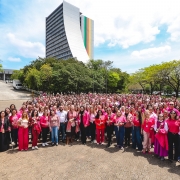 Uma fotografia colorida em plano geral e perspetiva em contra-plong&eacute;e suave (vista de &acirc;ngulo elevado) capta uma grande foto coletiva de servidores p&uacute;blicos estaduais reunidos em apoio &agrave; campanha do Outubro Rosa no p&aacute;tio do Centro Administrativo Fernando Ferrari (CAFF), em Porto Alegre.

**Os Servidores e o Movimento Outubro Rosa (em primeiro e meio plano):**

* Em primeiro plano e no centro da composi&ccedil;&atilde;o, uma multid&atilde;o formada por dezenas de servidores p&uacute;blicos pose alinhada e sorridente para a fotografia oficial.


* A grande maioria dos participantes veste pe&ccedil;as de vestu&aacute;rio em tons de cor-de-rosa (t-shirts, blusas, casacos, blazers e cal&ccedil;as) e muitos seguram pequenas rosas cor-de-rosa ou brancas nas m&atilde;os.


* No centro do grupo da frente, destaca-se a secret&aacute;ria estadual da Sa&uacute;de, Arita Bergmann, acompanhada por gestores e lideran&ccedil;as da pasta.


* O grupo encontra-se reunido sobre o pavimento intertravado de bet&atilde;o (paver) de uma grande esplanada exterior.



**O Edif&iacute;cio do CAFF e o Entorno (ao fundo):**

* Ao fundo, no centro superior da imagem, ergue-se o ic&oacute;nico edif&iacute;cio principal do Centro Administrativo Fernando Ferrari (CAFF), com a sua arquitetura vertical curva caracter&iacute;stica e um painel de grandes dimens&otilde;es nas cores da bandeira do Rio Grande do Sul (verde, vermelho e amarelo) verticais na sua fachada.


* A ladear o edif&iacute;cio e em redor da esplanada, observam-se &aacute;rvores frondosas com folhagem verde intensa.


* &Agrave; esquerda, avista-se uma rampa de acesso ao complexo e ve&iacute;culos estacionados.


* O c&eacute;u apresenta-se num tom azul-claro com nuvens brancas e esparsas, sob ilumina&ccedil;&atilde;o solar direta de dia claro.