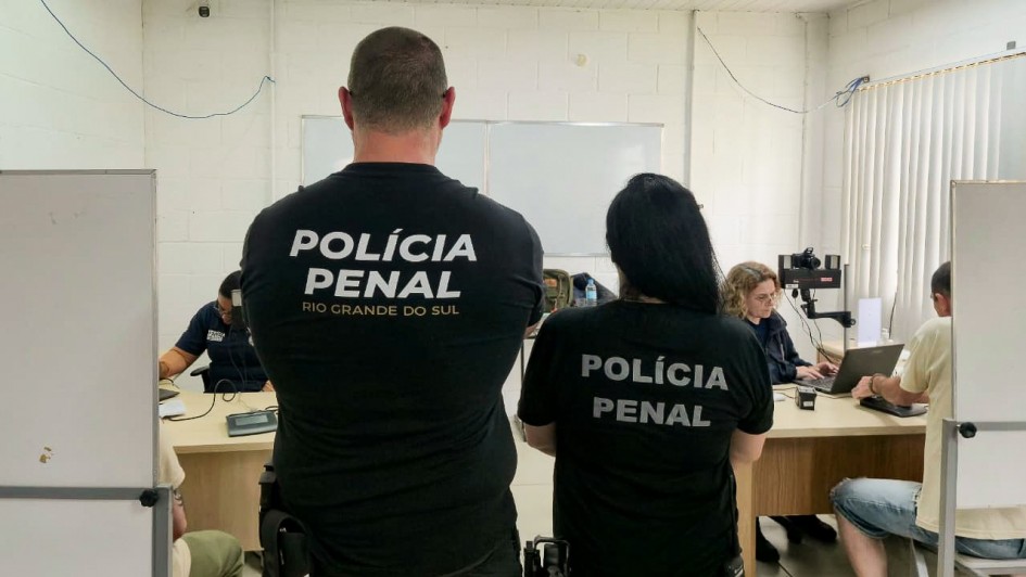 Foto mostra dois policiais penais, de costa, com camisetas pretas da Pol&iacute;cia Penal, na frente de uma sala onde atendentes est&atilde;o coletando os dados dos detentos. 