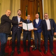 Grupo de pessoas posa sobre o palco exibindo um documento aberto para a c&acirc;mera, durante uma cerim&ocirc;nia oficial realizada em um sal&atilde;o hist&oacute;rico com bandeiras ao fundo e tapete vermelho.