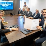 Uma fotografia colorida em plano m&eacute;dio e perspectiva em &acirc;ngulo levemente elevado capta o governador Eduardo Leite e gestores estaduais durante o ato de assinatura do decreto de institui&ccedil;&atilde;o do Programa Avan&ccedil;ar Deep Tech.

**As Autoridades e a Assinatura (em primeiro plano):**

* Em primeiro plano, &agrave; direita, o governador Eduardo Leite sorri para a c&acirc;mara enquanto segura com ambas as m&atilde;os uma pasta com o decreto oficial assinado. Ele veste um blazer azul-marinho sobre camisola de gola redonda clara, fita/cord&atilde;o institucional ao pesco&ccedil;o e rel&oacute;gio digital com mostrador verde no pulso esquerdo.
* &Agrave; esquerda, em primeiro plano, um gestor sorri virado para a c&acirc;mara, trajando blazer escuro e camisa social branca de colarinho aberto, com o bra&ccedil;o esquerdo apoiado sobre uma pasta de couro na mesa.
* Ambos est&atilde;o sentados em cadeiras de escrit&oacute;rio estofadas em tom cinzento-claro nas extremidades de uma grande mesa de reuni&otilde;es retangular de madeira escura.

**A Mesa de Reuni&atilde;o e o Painel (ao fundo):**

* Ao longo do lado oposto da mesa, alinham-se cerca de meia dezena de secret&aacute;rios, diretores e t&eacute;cnicos em trajes formais (fatos, blazers e gravatas), acompanhando atentamente o ato de assinatura.
* Ao fundo, &agrave; esquerda, sobressai um ecr&atilde; de televis&atilde;o fixado numa parede com pain&eacute;is de madeira ripada vertical. O ecr&atilde; exibe a apresenta&ccedil;&atilde;o oficial com o cabe&ccedil;alho **"ASSINATURA DE DECRETO"** e o t&iacute;tulo principal em letras brancas: **"Programa Avan&ccedil;ar Deep Tech"** sobre fundo azul tecnol&oacute;gico.
* Ao fundo e ao centro, um assessor em p&eacute; regista o momento com um smartphone.
* O ambiente conta com ilumina&ccedil;&atilde;o interior clara e uniforme, transmitindo um clima solene e institucional.