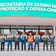 Des pessoas estao lado a lado na frente da entrada de um pr&eacute;dio onde tem a inscri&ccedil;&atilde;o Secretaria de Estado da Prote&ccedil;&atilde;o e Defesa Civil