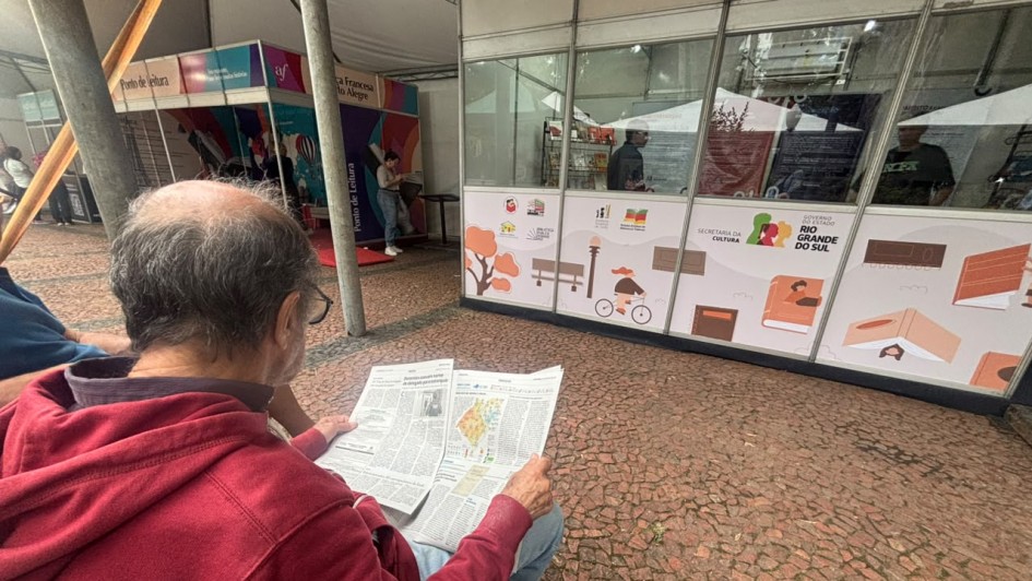 Imagem mostra um homem em primeiro plano lendo um folheto sentado em frente a um estante da Secretaria da Cultura do Rio Grande do Sul na Feira do Livro de Porto Alegre 